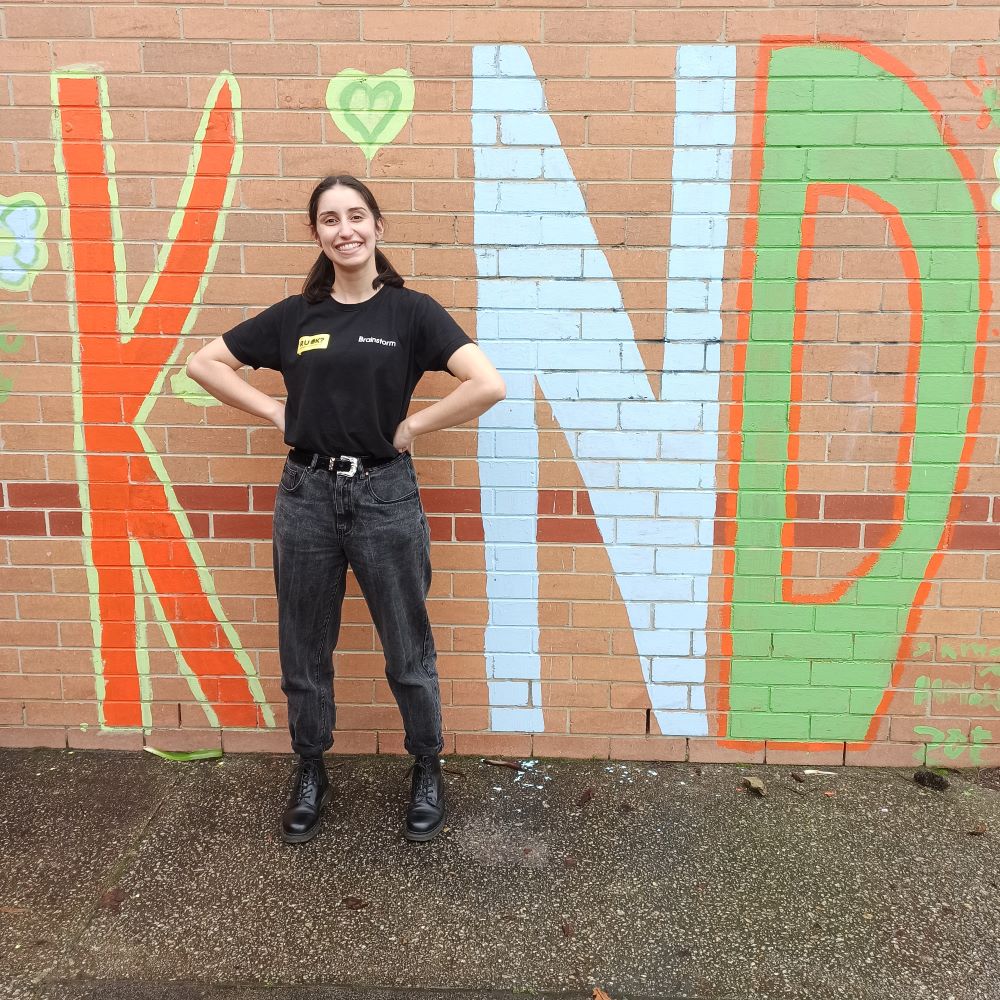 A Brainstorm Productions actor standing in front of a school mural that says 'Kind' and she is being the letter 'I'.