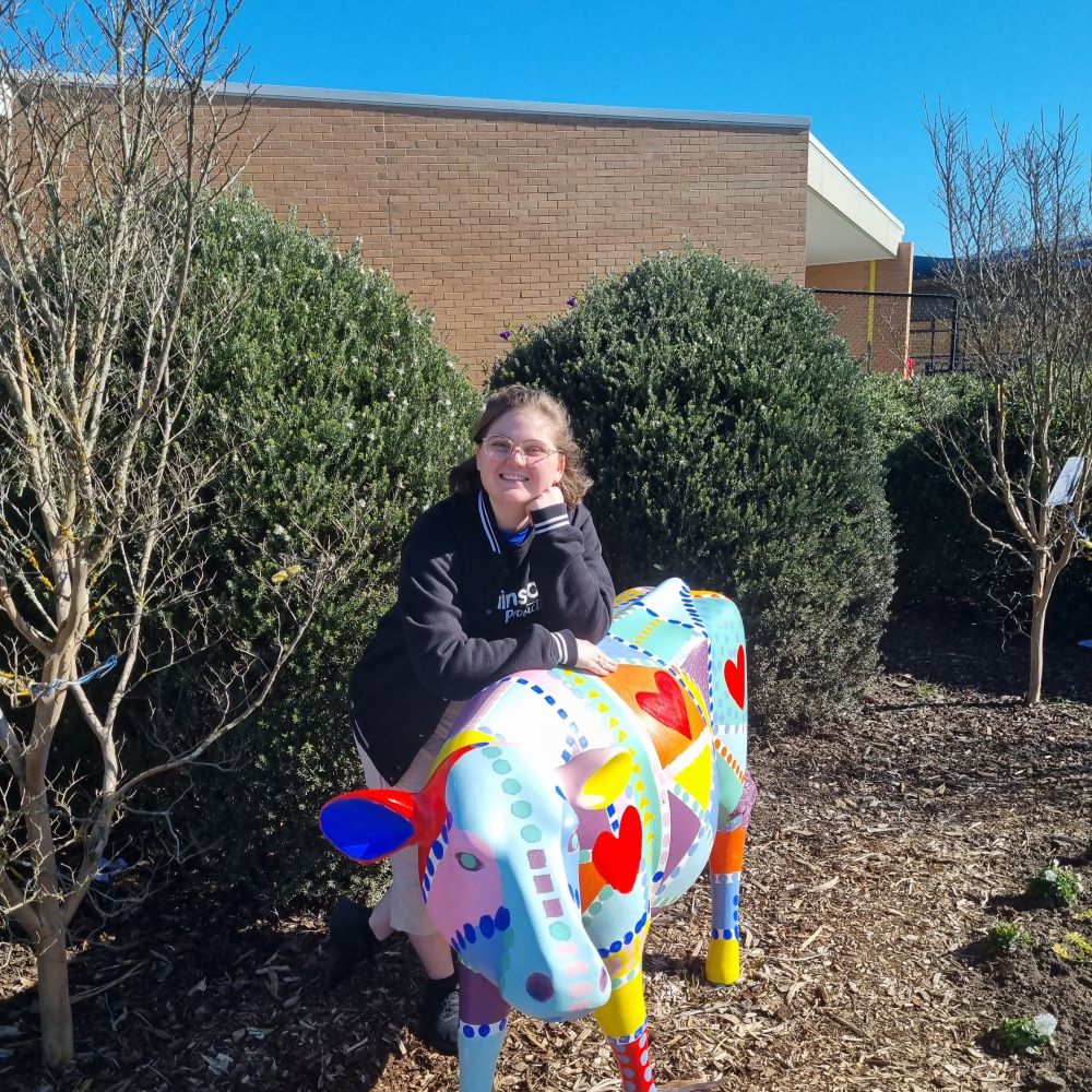 A Brainstorm Productions actor smiling and leaning on a colourfully painted cow sculpture at a regional Victorian primary school