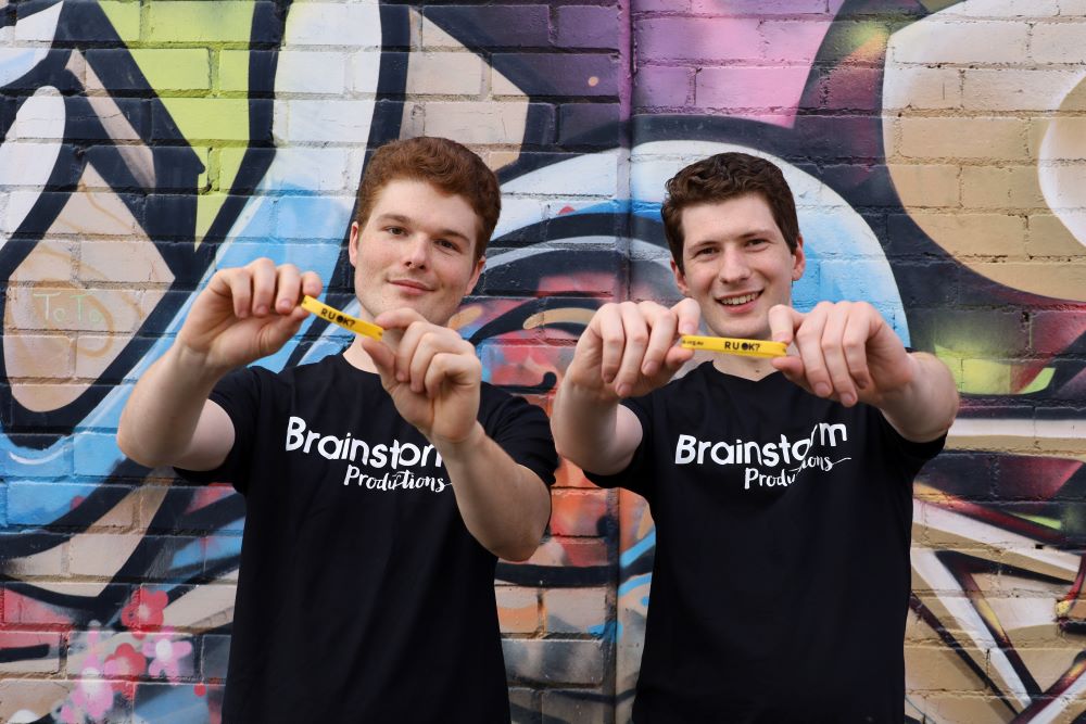 Two Brainstorm Productions actors are holding R U OK? wristbands towards the camera and standing in front of a colourful street art wall