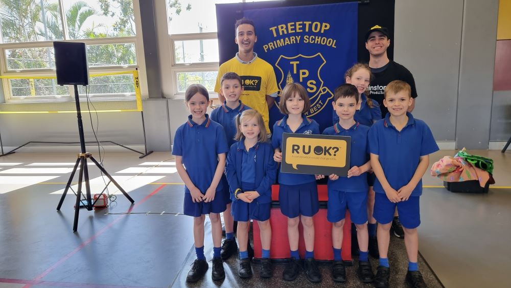 Two Brainstorm Productions actors, Jake and Raphael, are smiling with a group of QLD primary school students on R U OK?Day 2023