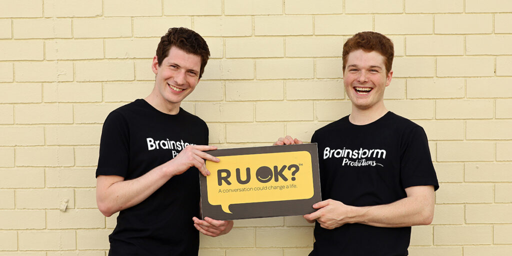 Two Brainstorm Productions actors wearing company shirts are holding an R U OK? sign and smiling for the camera, standing in front of a yellow wall