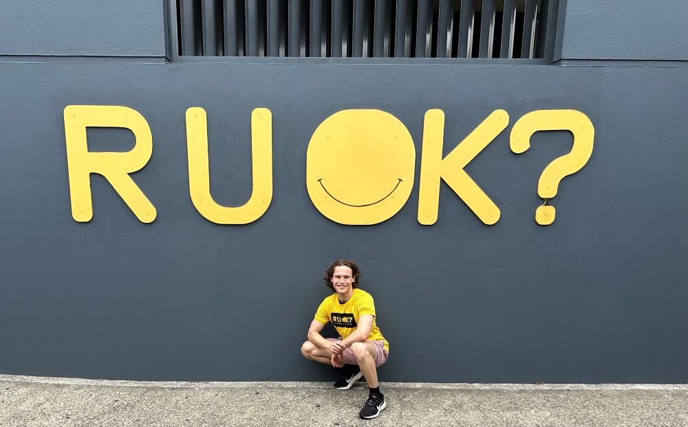 Brainstorm Productions actor Bailey is siling for the camera in front of an R U OK? sign and wearing an R U OK? t-shirt