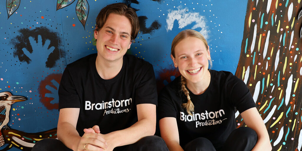 Two Brainstorm Productions actors, members of the 2025 theatre in education team, are smiling and sitting in front of a colourful painted wall.